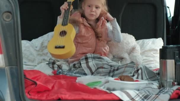 Hermosa niña sentada en un baúl abierto de un coche en la orilla del río del mar jugando en un ukelele amarillo — Vídeo de stock