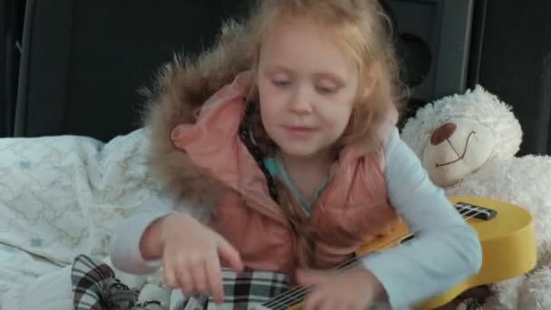 Hermosa niña sentada en un baúl abierto de un coche en la orilla del río del mar jugando en un ukelele amarillo — Vídeos de Stock