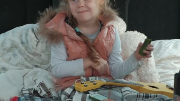 Schönes kleines Mädchen, das im offenen Kofferraum eines Autos am Ufer des Meeres sitzt und auf einer gelben Ukulele spielt — Stockvideo