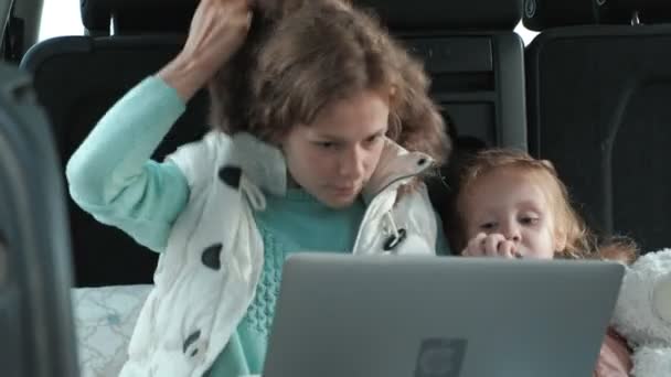 Beautiful young woman and her little daughter are sitting in the open trunk of a car on the river bank of the sea enjoying a laptop — Stock Video
