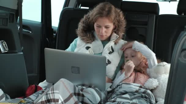 Mulher bonita e sua filhinha estão sentados no tronco aberto de um carro na margem do rio do mar desfrutando de um laptop — Vídeo de Stock