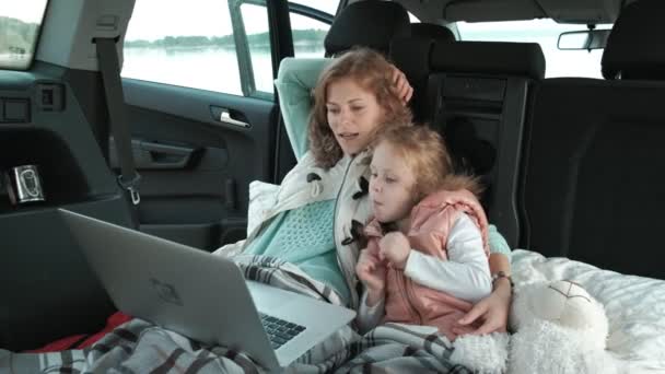 Beautiful young woman and her little daughter are sitting in the open trunk of a car on the river bank of the sea enjoying a laptop — Stock Video