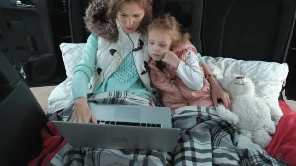 Красива молода жінка і її маленька дочка сидять у відкритому багажнику автомобіля на березі річки моря, насолоджуючись ноутбуком — стокове відео