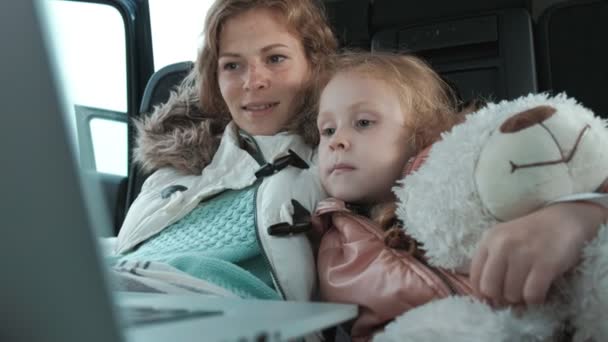 Bella giovane donna e la sua piccola figlia sono seduti nel bagagliaio aperto di una macchina sulla riva del mare godendo di un computer portatile — Video Stock
