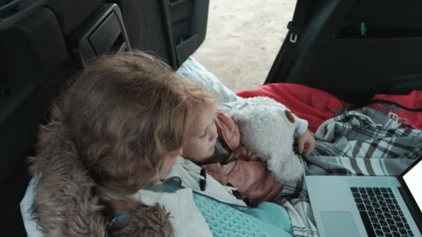 Mooie jonge vrouw en haar dochtertje zitten in de open kofferbak van een auto op de oever van de zee genieten van een laptop — Stockvideo