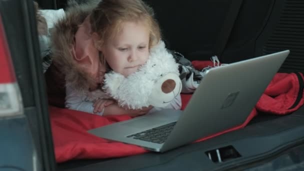 Mulher bonita e sua filhinha estão sentados no tronco aberto de um carro na margem do rio do mar desfrutando de um laptop — Vídeo de Stock