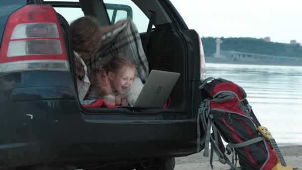 Mulher bonita e sua filhinha estão sentados no tronco aberto de um carro na margem do rio do mar desfrutando de um laptop — Vídeo de Stock