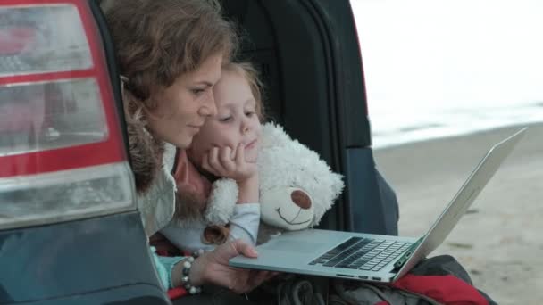 아름 다운 젊은 여자와 그녀의 작은 딸 노트북을 즐기는 바다의 강둑에는 자동차의 열려있는 트렁크에 앉아 있다 — 비디오