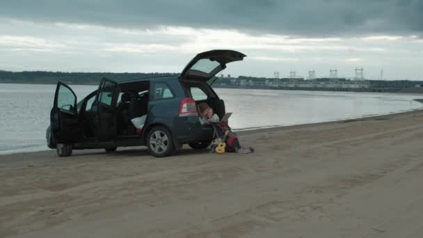 ノート パソコンを楽しんで海の川の土手に車の開いているトランクの美しい若い女性と彼女の小さな娘が座っています。 — ストック動画