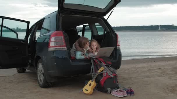 Hermosa joven y su hija pequeña están sentados en el maletero abierto de un coche en la orilla del río del mar disfrutando de un ordenador portátil — Vídeo de stock