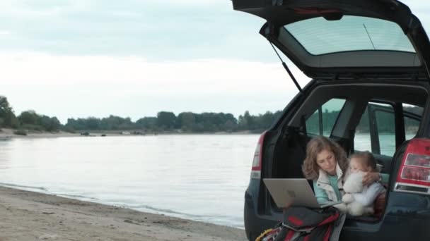 Hermosa joven y su hija pequeña están sentados en el maletero abierto de un coche en la orilla del río del mar disfrutando de un ordenador portátil — Vídeos de Stock