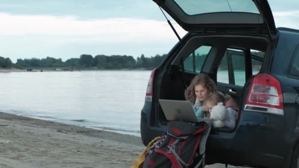 Красива молода жінка і її маленька дочка сидять у відкритому багажнику автомобіля на березі річки моря, насолоджуючись ноутбуком — стокове відео
