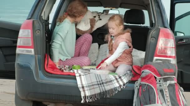 Hermosa joven mujer y su hija pequeña están sentados en el tronco abierto de un coche en la orilla de un río del mar comiendo frutas y beber té de un termo — Vídeos de Stock