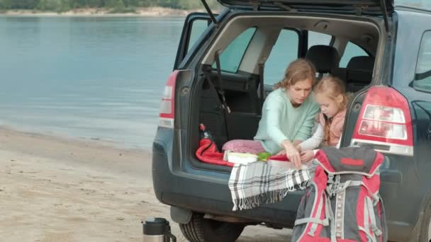 Mulher nova bonita e sua filha pequena estão sentados no tronco aberto de um carro na margem de um rio do mar comendo frutas e bebendo chá de uma garrafa térmica — Vídeo de Stock