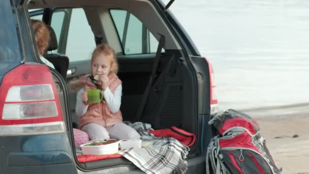 Schöne junge Frau und ihre kleine Tochter sitzen im offenen Kofferraum eines Autos am Ufer eines Meeres und essen Früchte und trinken Tee aus einer Thermoskanne — Stockvideo