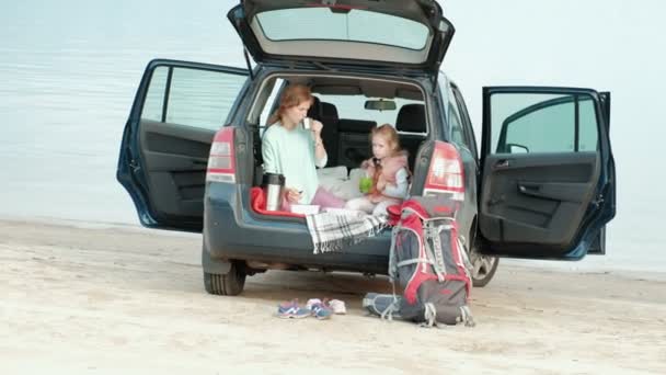 Piękna młoda kobieta i jej córeczka siedzą w otwartym bagażniku samochodu, na brzegu rzeki, morza, owoce do jedzenia i picia herbaty z termos — Wideo stockowe