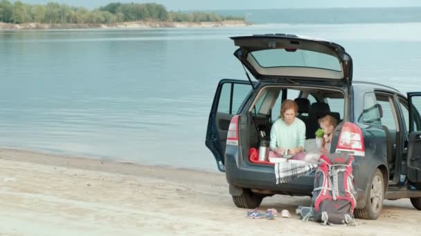 Beautiful young woman and her little daughter are sitting in the open trunk of a car on the bank of a river of the sea eating fruits and drinking tea from a thermos — Stock Video