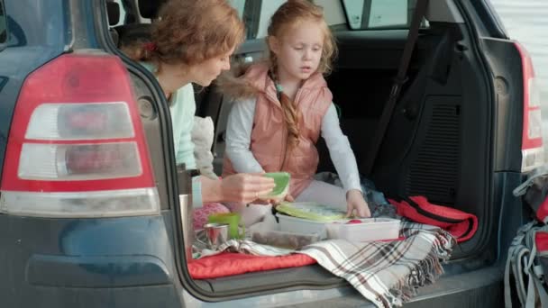 Hermosa joven mujer y su hija pequeña están sentados en el tronco abierto de un coche en la orilla de un río del mar comiendo frutas y beber té de un termo — Vídeos de Stock