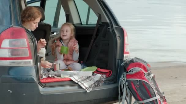 Mulher nova bonita e sua filha pequena estão sentados no tronco aberto de um carro na margem de um rio do mar comendo frutas e bebendo chá de uma garrafa térmica — Vídeo de Stock