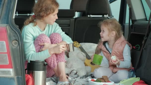 Schöne junge Frau und ihre kleine Tochter sitzen im offenen Kofferraum eines Autos am Ufer eines Meeres und essen Früchte und trinken Tee aus einer Thermoskanne — Stockvideo