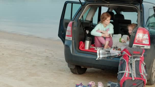 Mooie jonge vrouw en haar dochtertje zitten in de open kofferbak van een auto op de oever van een rivier in de zee vruchten eten en het drinken van thee uit een thermos — Stockvideo