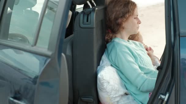 Hermosa joven mujer y su hija pequeña están sentados en el tronco abierto de un coche en la orilla de un río del mar comiendo frutas y beber té de un termo — Vídeos de Stock