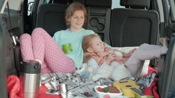 Beautiful young woman and her little daughter are sitting in the open trunk of a car on the bank of a river of the sea eating fruits and drinking tea from a thermos — Stock Video