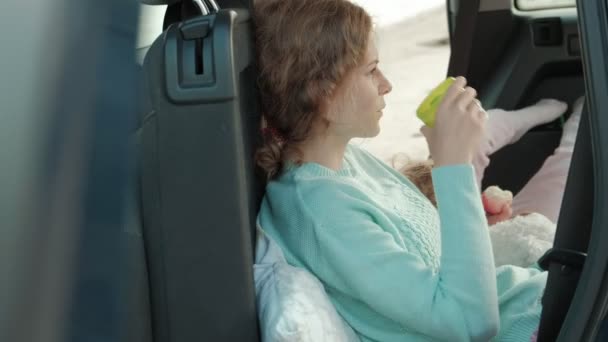 Hermosa joven mujer y su hija pequeña están sentados en el tronco abierto de un coche en la orilla de un río del mar comiendo frutas y beber té de un termo — Vídeos de Stock