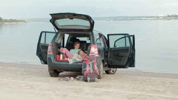 Vacker ung kvinna och hennes lilla dotter sitter i öppna bagageluckan på en bil på stranden av en flod av havet äta frukt och dricka te från en termos — Stockvideo