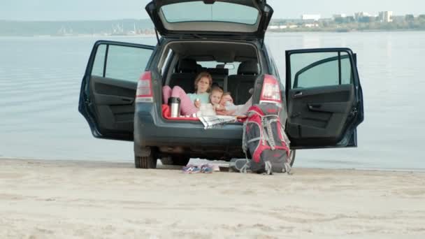 Hermosa joven mujer y su hija pequeña están sentados en el tronco abierto de un coche en la orilla de un río del mar comiendo frutas y beber té de un termo — Vídeo de stock