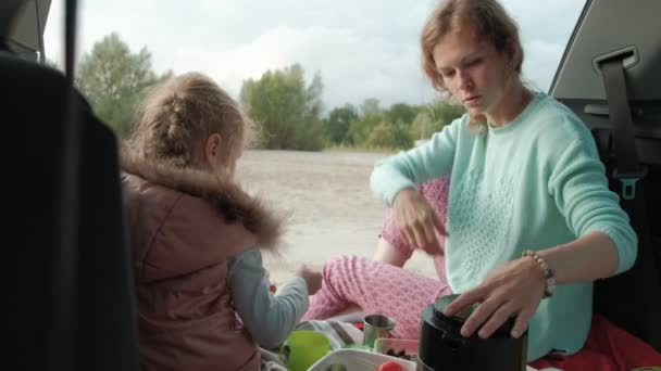 Mooie jonge vrouw en haar dochtertje zitten in de open kofferbak van een auto op de oever van een rivier in de zee vruchten eten en het drinken van thee uit een thermos — Stockvideo