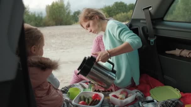 美しい若い女性と彼女の小さな娘は果物を食べると、ポットからお茶を飲んで海の川の銀行の車の開いているトランクに座っています。 — ストック動画