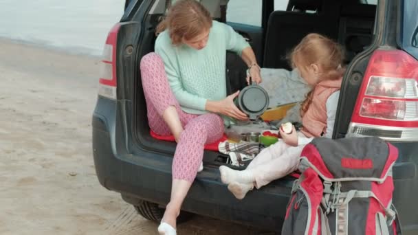 Mooie jonge vrouw en haar dochtertje zitten in de open kofferbak van een auto op de oever van een rivier in de zee vruchten eten en het drinken van thee uit een thermos — Stockvideo