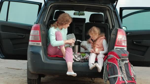 Schöne junge Frau und ihre kleine Tochter sitzen im offenen Kofferraum eines Autos am Ufer eines Meeres und essen Früchte und trinken Tee aus einer Thermoskanne — Stockvideo