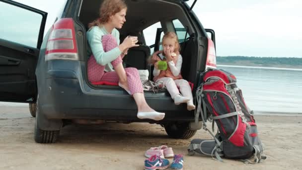 Mulher nova bonita e sua filha pequena estão sentados no tronco aberto de um carro na margem de um rio do mar comendo frutas e bebendo chá de uma garrafa térmica — Vídeo de Stock