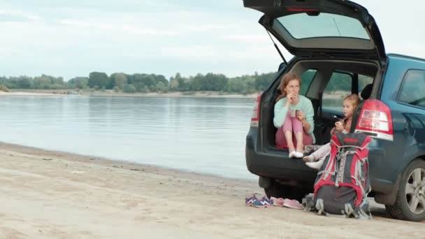 Красивая молодая женщина и ее маленькая дочь сидят в открытом багажнике автомобиля на берегу реки моря, едят фрукты и пьют чай из термоса — стоковое видео
