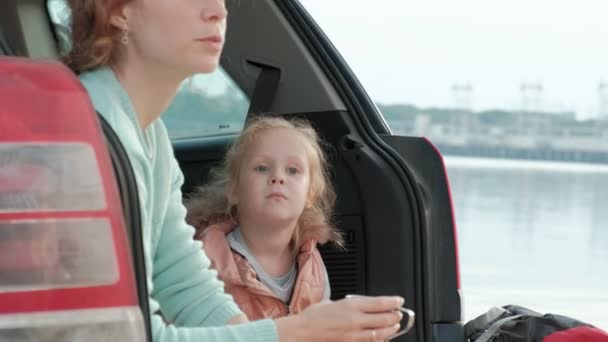 Hermosa joven mujer y su hija pequeña están sentados en el tronco abierto de un coche en la orilla de un río del mar comiendo frutas y beber té de un termo — Vídeos de Stock