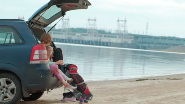 Mulher nova bonita e sua filha pequena estão sentados no tronco aberto de um carro na margem de um rio do mar comendo frutas e bebendo chá de uma garrafa térmica — Vídeo de Stock