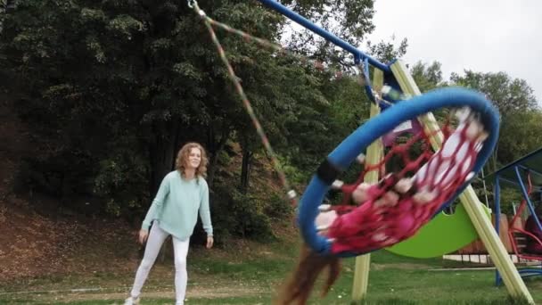 Een klein meisje in een roze jurk swingt op een ronde schommel in de speeltuin — Stockvideo