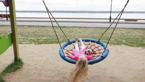 Una niña en un vestido rosa se balancea en un columpio redondo en el patio de recreo — Vídeos de Stock