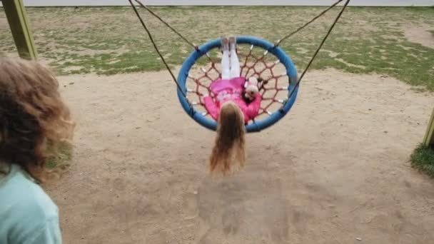 Маленька дівчинка в рожевому платті гойдається на круглих гойдалках на дитячому майданчику — стокове відео