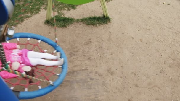 Una niña en un vestido rosa se balancea en un columpio redondo en el patio de recreo — Vídeos de Stock