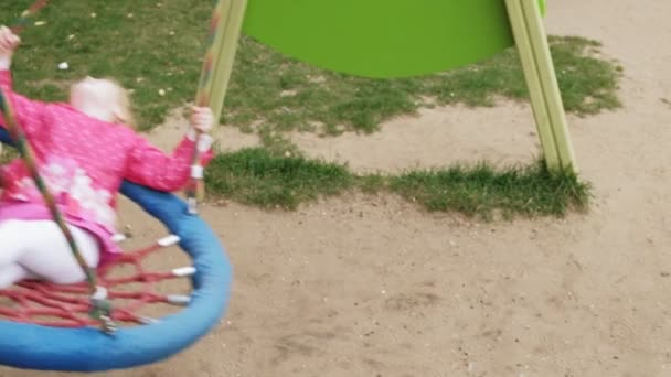 Una niña en un vestido rosa se balancea en un columpio redondo en el patio de recreo — Vídeo de stock