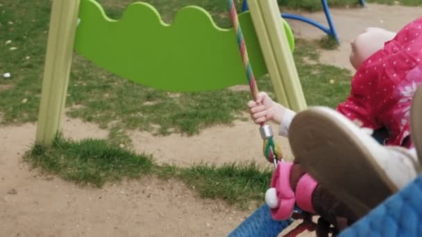 Una niña en un vestido rosa se balancea en un columpio redondo en el patio de recreo — Vídeos de Stock