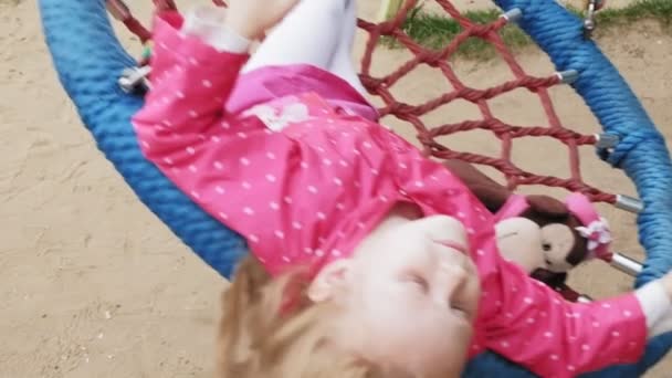 Una niña en un vestido rosa se balancea en un columpio redondo en el patio de recreo — Vídeos de Stock