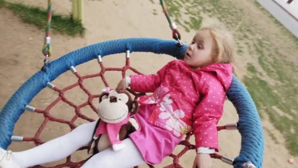 Ein kleines Mädchen im rosa Kleid schaukelt auf einer runden Schaukel auf dem Spielplatz — Stockvideo