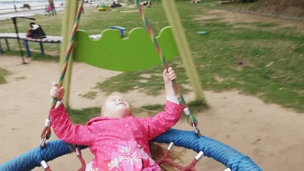 Uma menina em um vestido rosa balança em um balanço redondo no playground — Vídeo de Stock