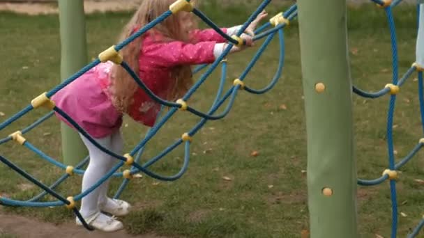 Egy kislány egy rózsaszín ruhát, és az anyja játszik a kötélhágcsót a játszótéren — Stock videók