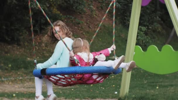 Uma menina em um vestido rosa e sua mãe balançando em um balanço redondo no playground — Vídeo de Stock