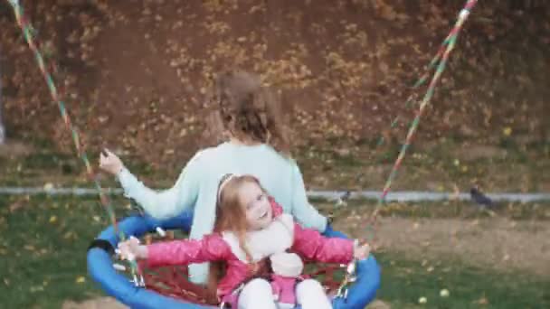 Uma menina em um vestido rosa e sua mãe balançando em um balanço redondo no playground — Vídeo de Stock